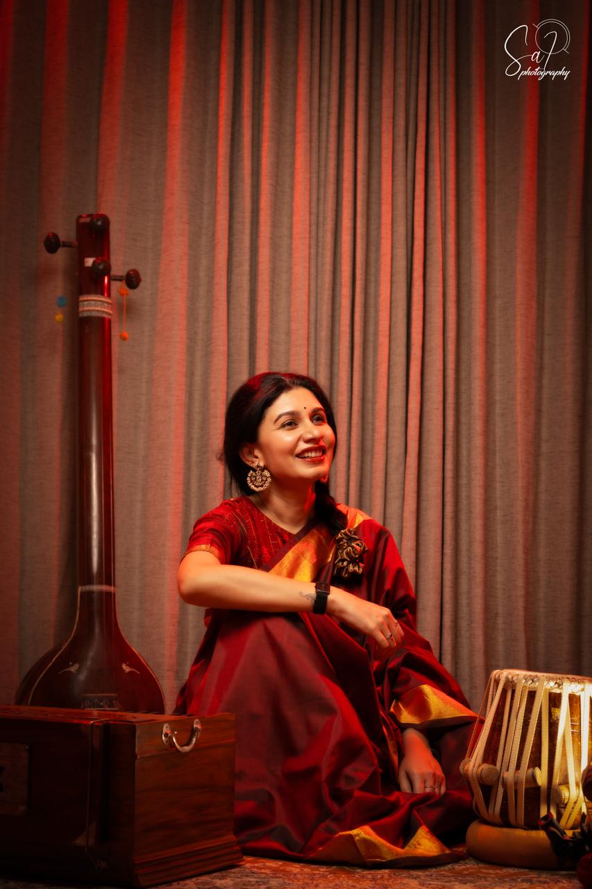 Harini Rao seated in a warm lamp-lit salon with tanpura and tabla, wearing a red and gold silk saree